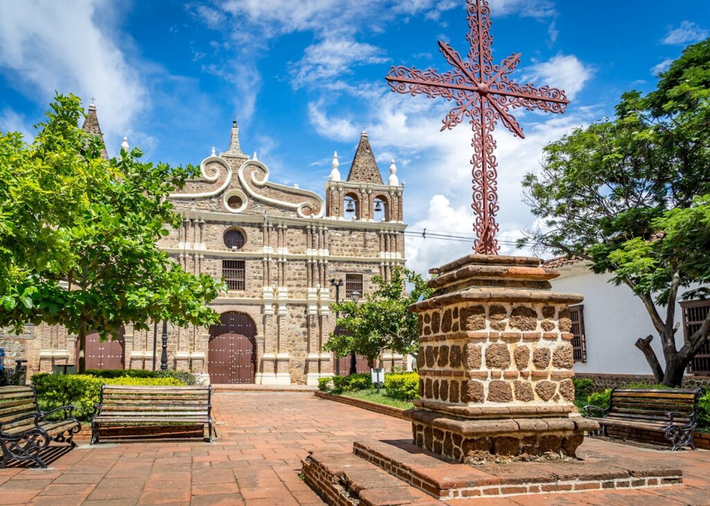 A sunlit view of Santa Fe de Antioquia showing colonial white houses with wooden balconies, cobblestone streets, and a central plaza shaded by palm trees, with the historic Cathedral of the Immaculate Conception rising in the background under a clear blue sky.